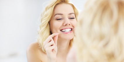 Junge, blonde Frau säubert ihre Zähne mit Zahnseide im Spiegel